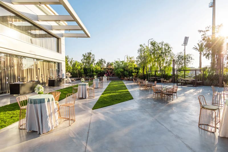 Terrasse dallée près d'un bâtiment moderne avec tables de cocktail, bande de pelouse et soleil bas à travers les arbres