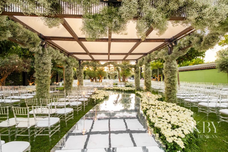 Wooden pergola with columns covered in white flowers over a mirrored aisle, with clear chairs and white flower beds