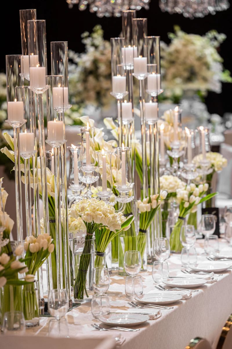 Table dressée avec hauts photophores en verre, bougies crème et fleurs blanches à hauteurs variées