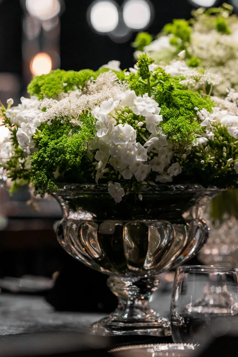 Coupe en verre sur pied garnie d’une dense composition de fleurs blanches et vertes, lumières floues derrière