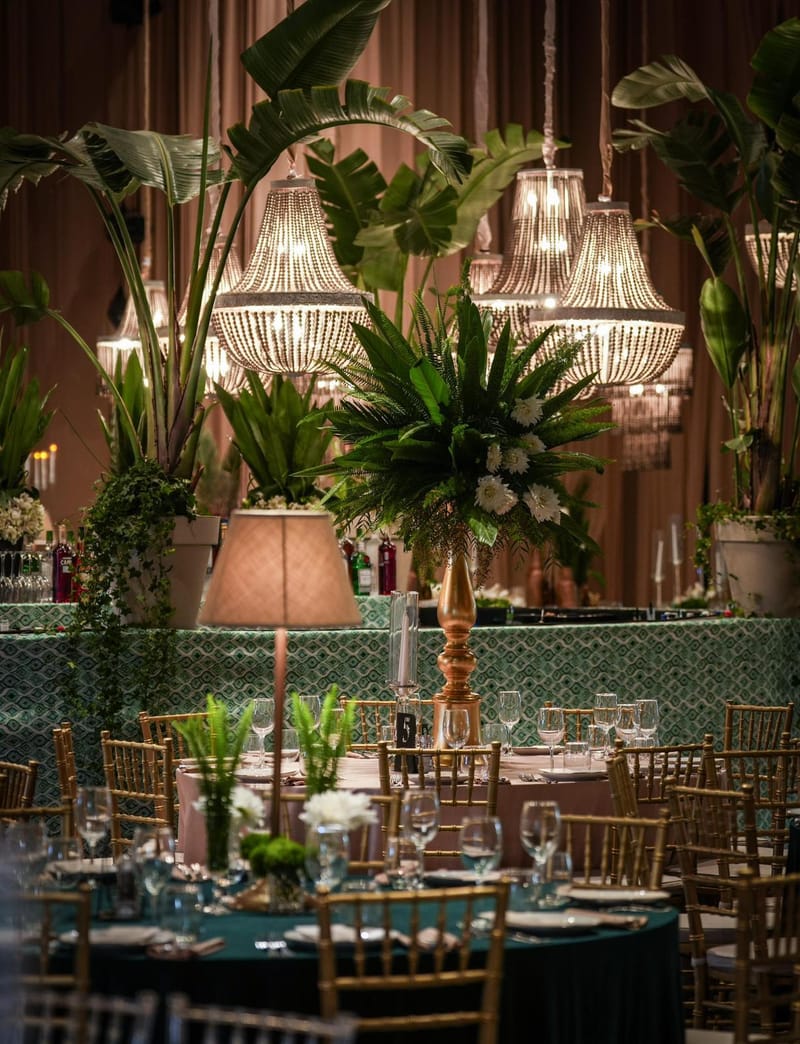 Large palm leaves beneath beaded chandeliers, a table lamp and round tables with gold chairs