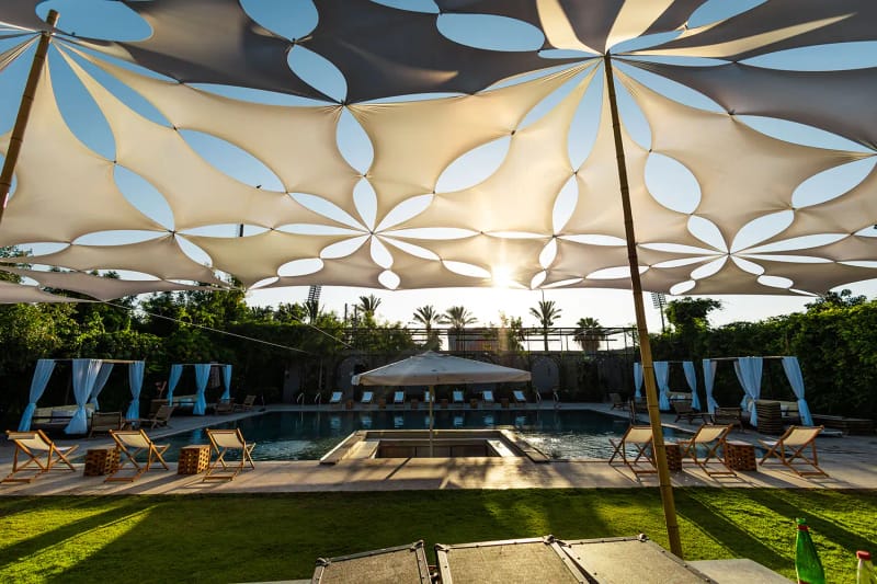 Voile d'ombrage crème aux découpes en forme de fleur au-dessus d'une piscine avec bains de soleil et cabines bleues