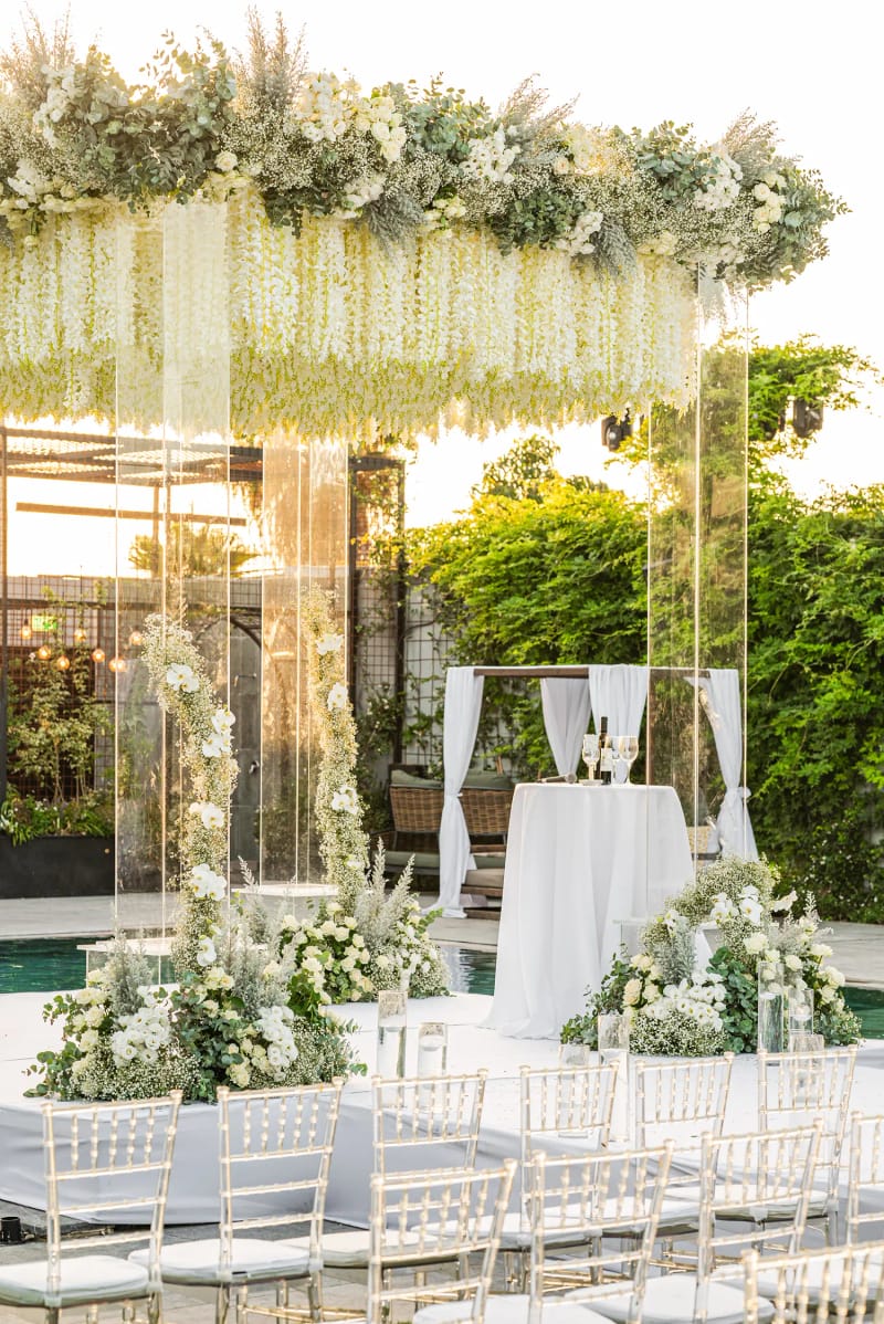 A chuppah with clear columns and a canopy of hanging white flowers, rows of white chairs and a draped pavilion by a pool