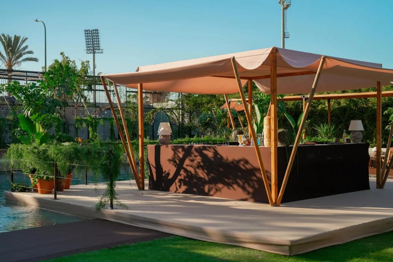 Terracotta fabric-roofed pavilion with a bar counter on a deck beside a pool and plants, under a blue sky