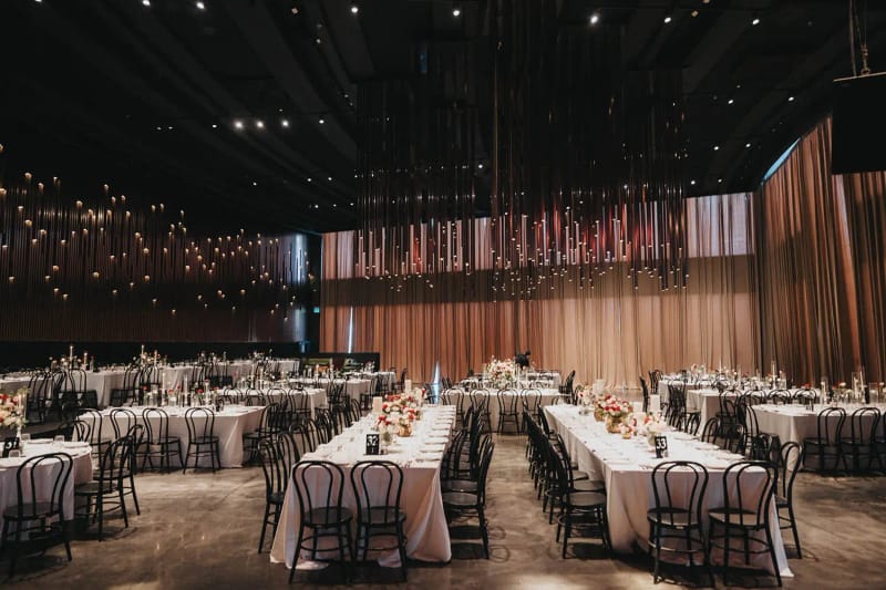 Vaste salle aux rangées de longues tables et chaises bistrot noires, rideau cuivré et fils lumineux suspendus au plafond