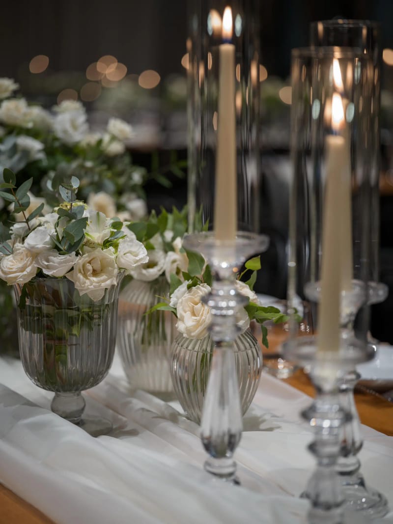 Taper candles in tall glass holders and glass vases with white flowers on a sheer table runner