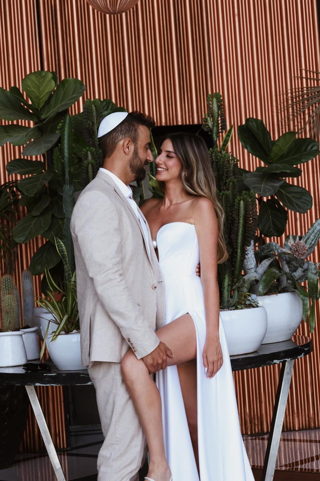 A couple embracing in front of a slatted wood wall and large potted plants, the groom wearing a kippah
