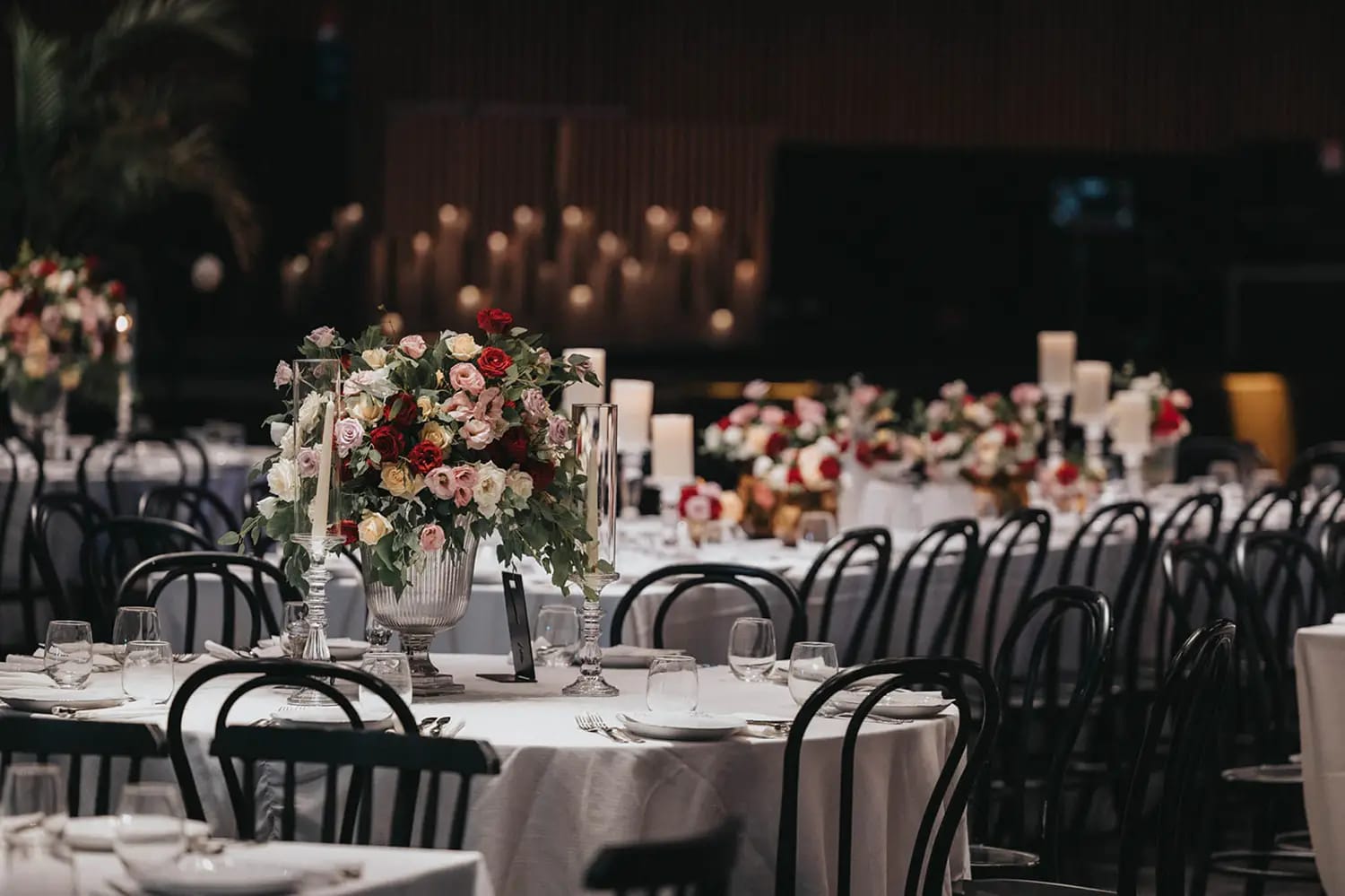 Tables rondes nappées de blanc avec chaises noires, centre de table rouge et rose et bougies dans une salle tamisée