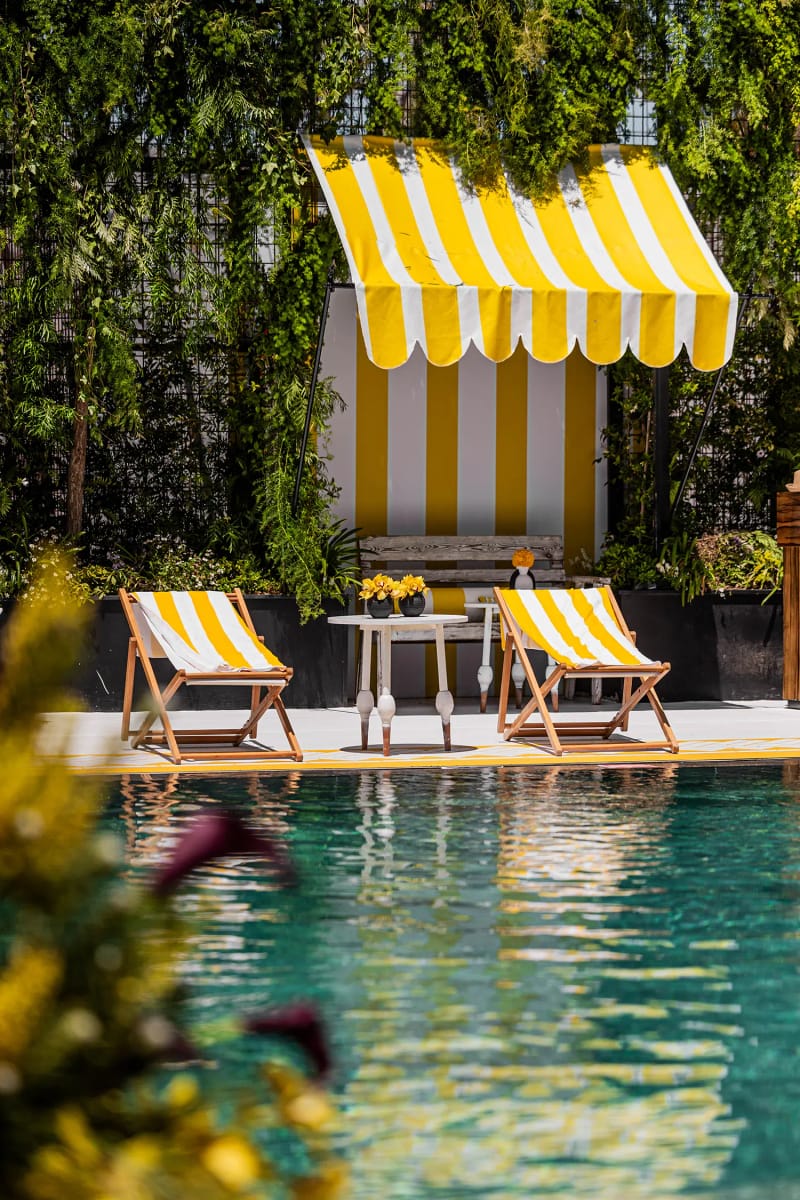 Yellow and white striped cabana with two striped deck chairs and a small table by the pool, in front of greenery