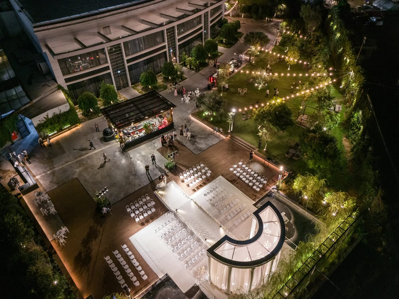 Vue aérienne de nuit d'un jardin à guirlandes lumineuses, estrade blanche de cérémonie entourée de chaises, bâtiment