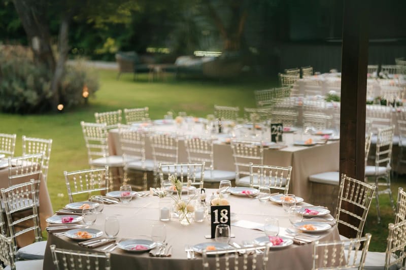 Table de banquet ronde dressée avec numéro de table parmi des chaises transparentes, longues tables sur la pelouse au fond