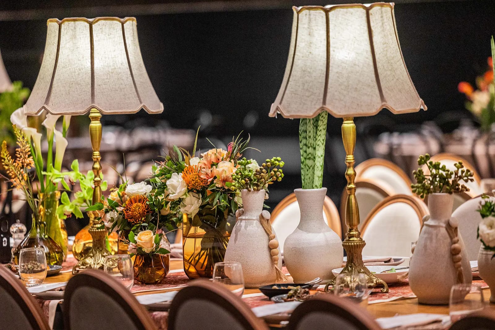 Longue table avec lampes à abat-jour de tissu, vases blancs et fleurs orange, chaises à dossier rond au premier plan