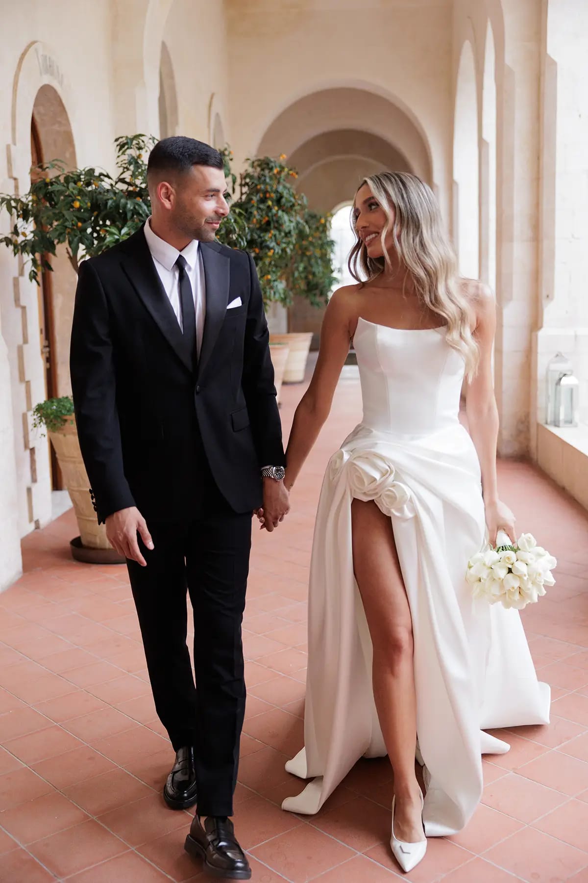 Un couple main dans la main dans une galerie voûtée en pierre au sol en terre cuite, la mariée tenant un bouquet blanc