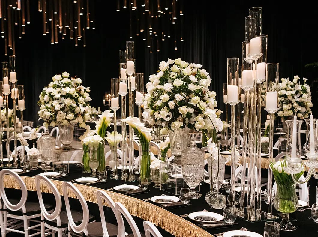 Table de banquet noire courbe au Shulhan, roses blanches, hauts photophores transparents et lumières dorées suspendues