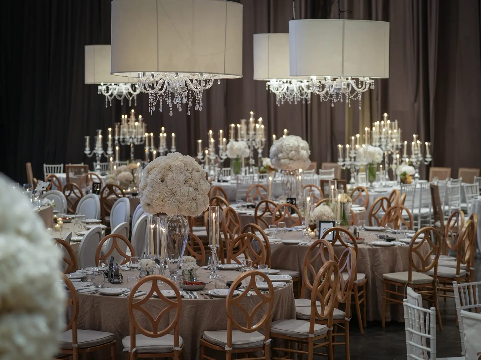Tables rondes avec chaises en bois doré, suspensions cylindriques et lustres, hauts chandeliers et boules de fleurs blanches