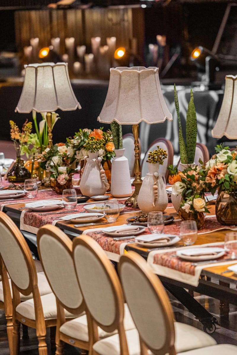 Table en bois avec lampes, fleurs et chaises à dossier arrondi, petites lumières suspendues dans la salle sombre au fond