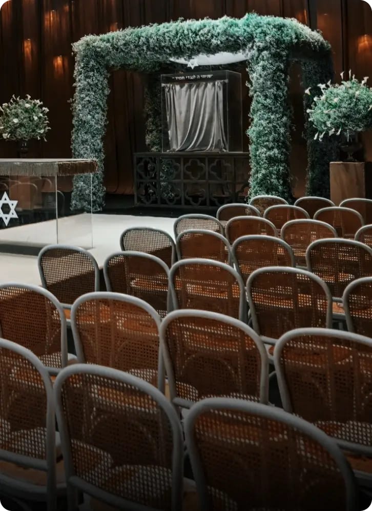 Une houppa couverte de feuillage vert pâle au-dessus d'une plateforme blanche, face à des rangées de chaises cannées