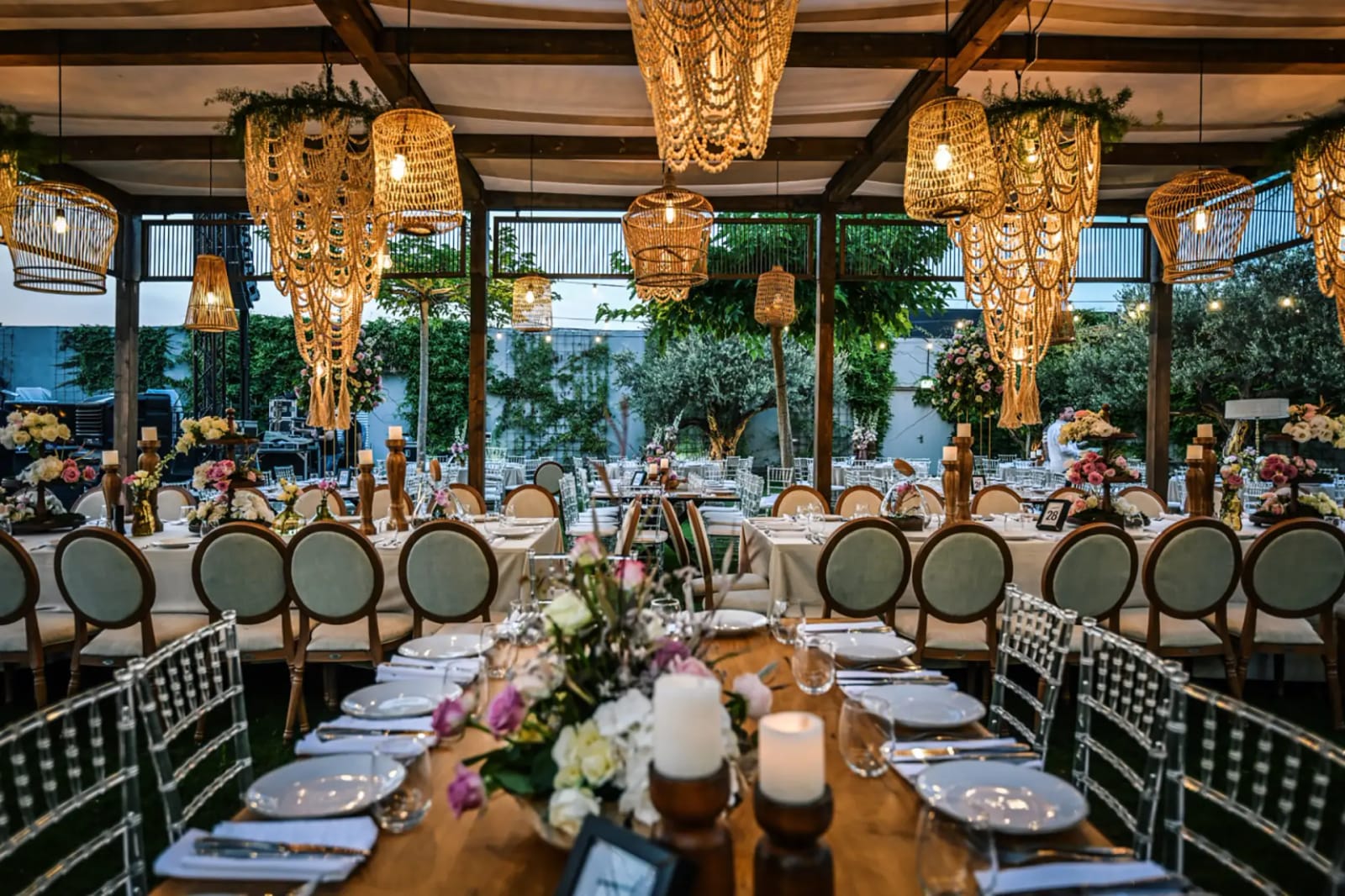 Long wooden table under a pergola with round-back chairs, woven rattan pendant lamps, pink flowers and candles