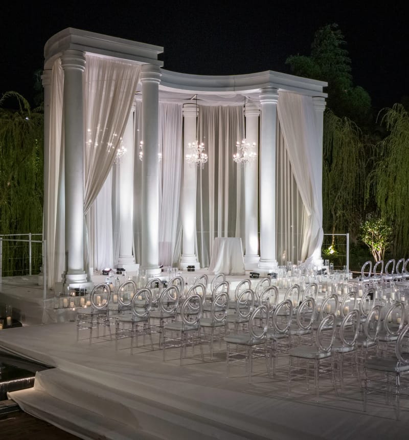 Pavillon blanc à colonnes et rideaux, rangées de chaises transparentes sur une estrade devant, entre des saules, de nuit