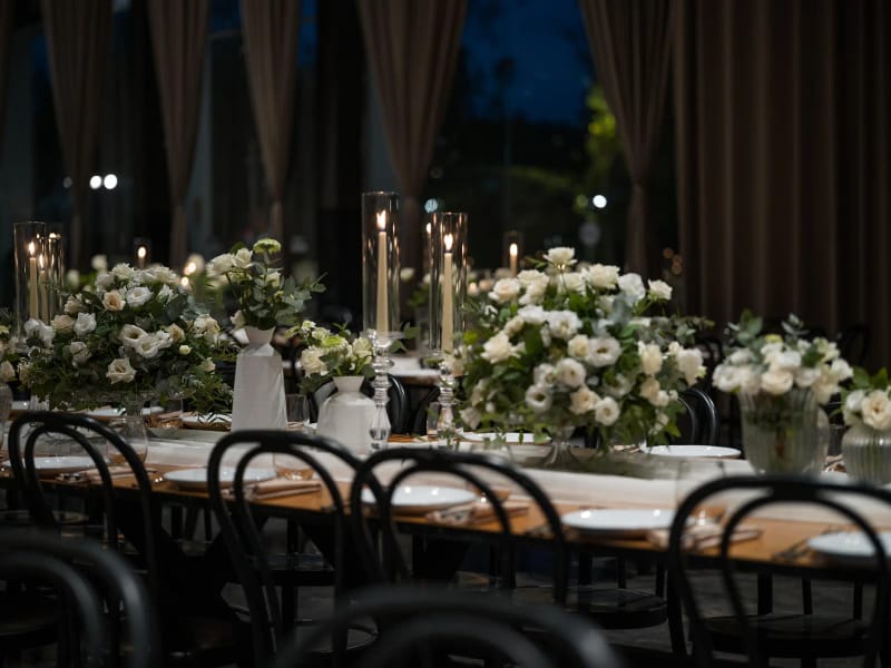 Long wooden table with vases of white flowers, tall candles and a white runner before curtains and a window