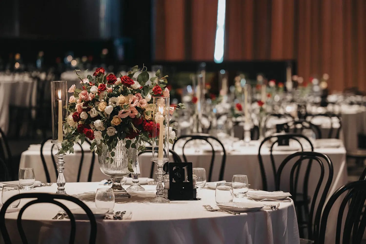Table ronde avec grand centre de table rouge et rose dans un vase de verre sur pied, chaises noires et rideaux bruns derrière