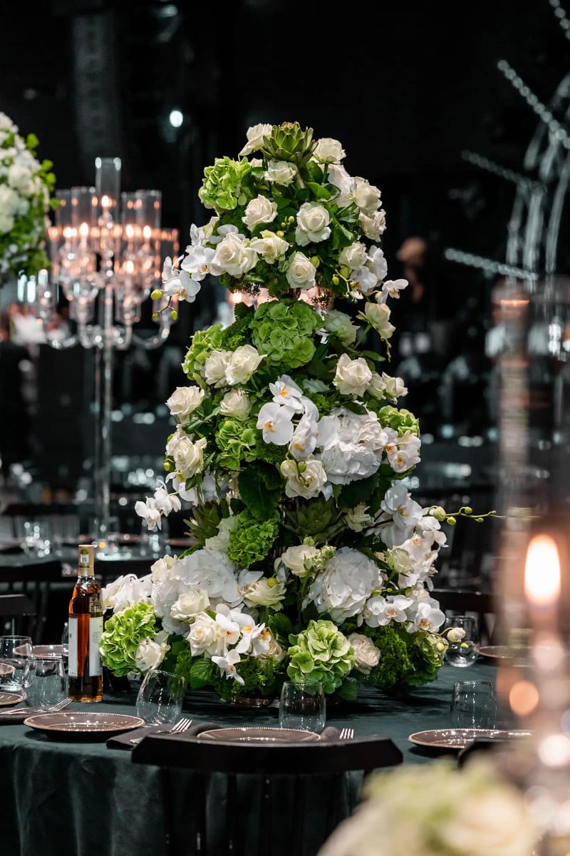 Haute tour de roses blanches et de fleurs vertes sur une table vert foncé, avec un candélabre allumé derrière