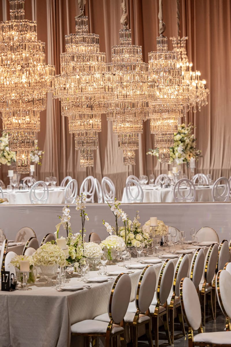 Lustres en cristal étagés au-dessus d’une longue table fleurie de blanc, chaises à dossier rond cerclé d’or