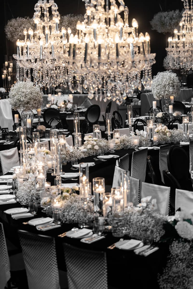 Crystal chandeliers above a long black table lined with candles in glass cylinders and white baby's breath