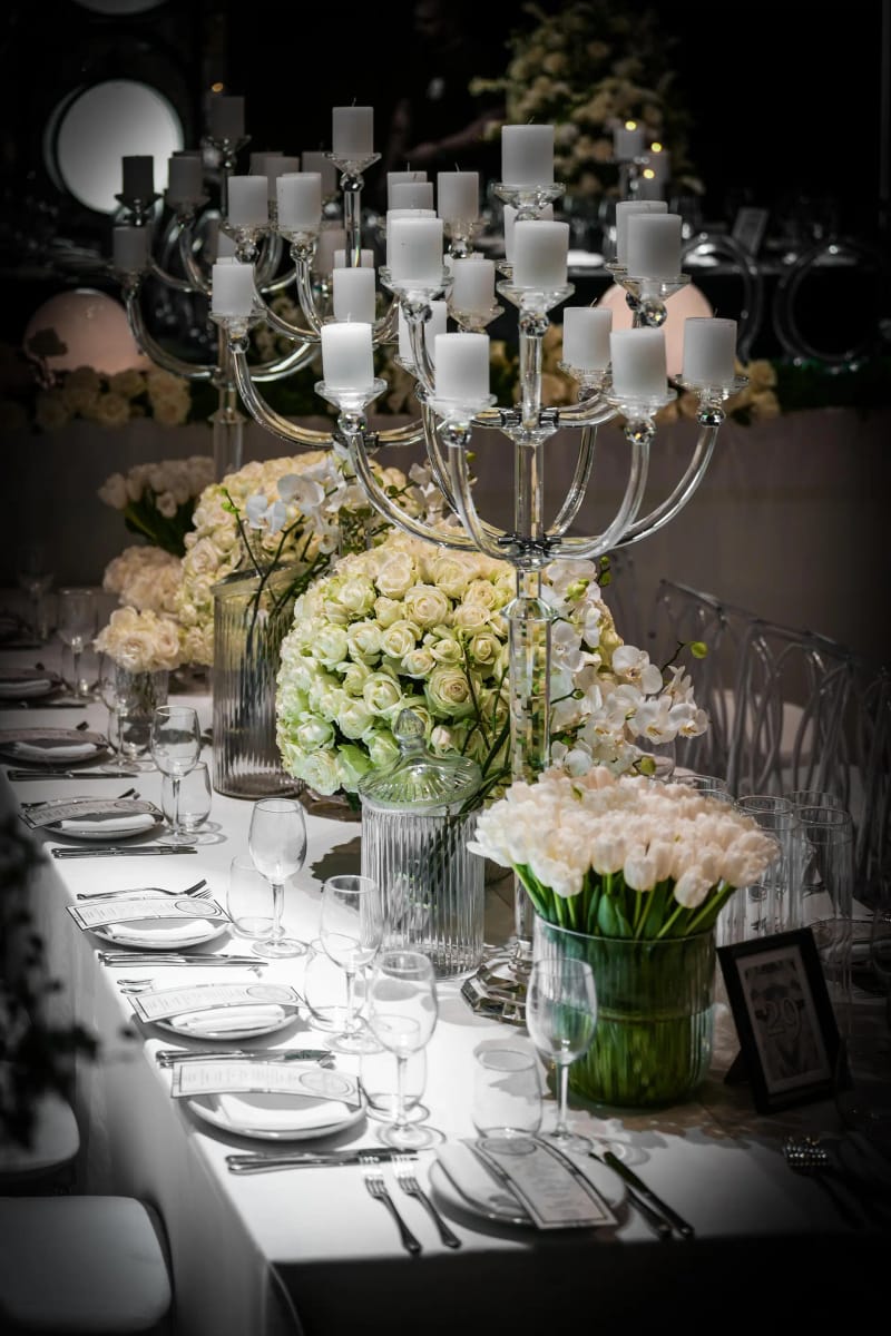 Longue table dressée avec un chandelier argenté à bougies blanches, roses et tulipes blanches dans des vases en verre