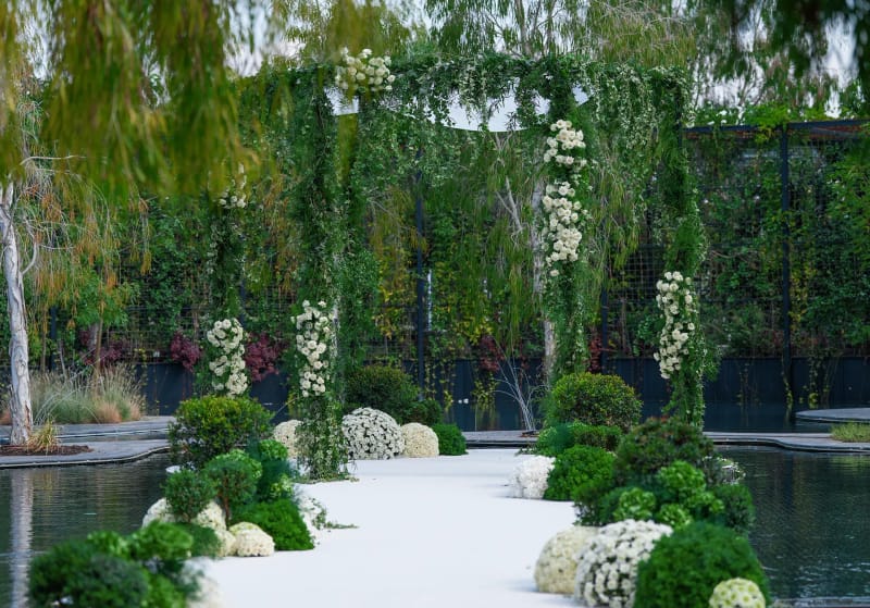 White platform at the pond edge with round shrubs and white flowers, and a greenery-wrapped canopy among willow trees
