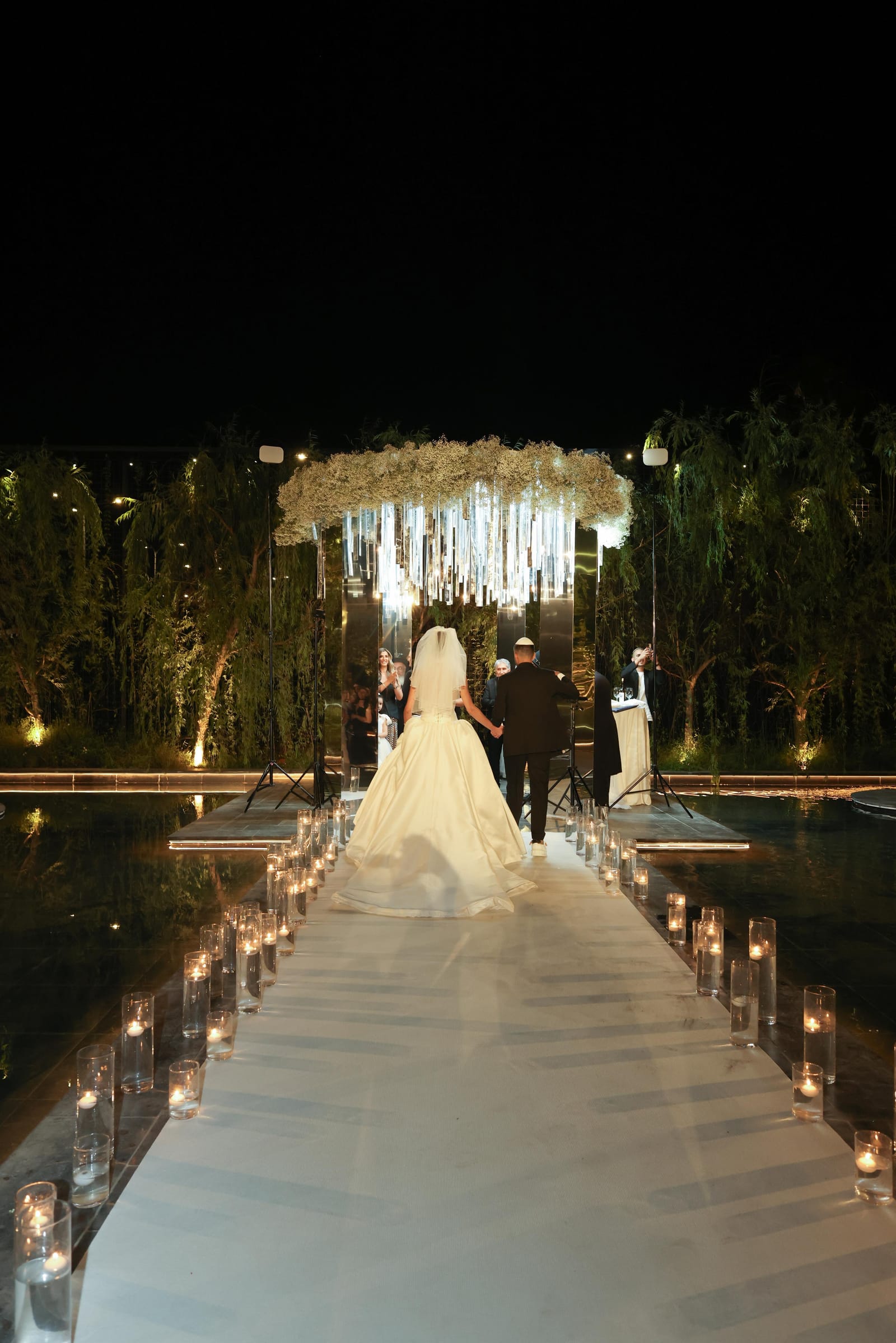 A bride in a gown with a long train walking down a candle-lined aisle over water toward a floral chuppah at night