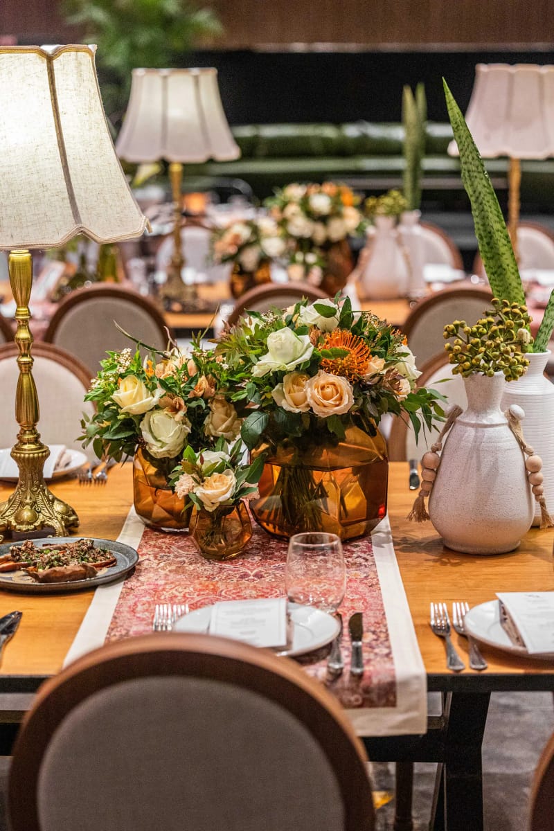Table avec lampe, fleurs orange et crème dans des vases blancs, chemin de table à motifs et assiettes, chaises à dossier rond