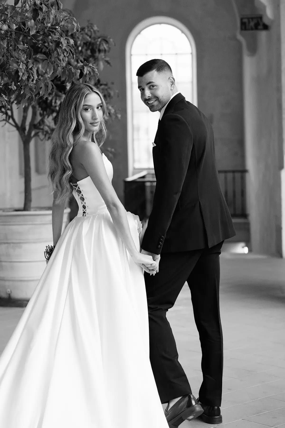 Image en noir et blanc d'un couple main dans la main dans un intérieur de pierre à fenêtre en arc, la mariée se retournant