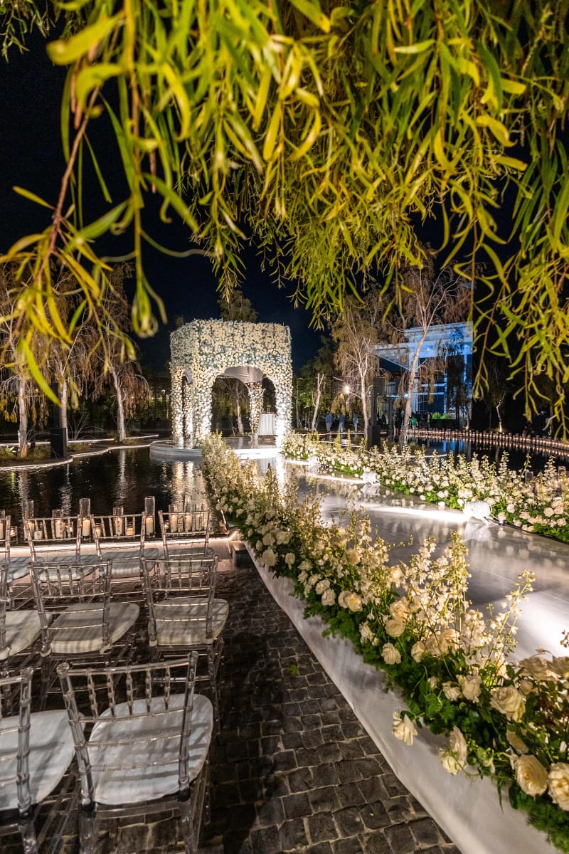 Arche de fleurs blanches au bout d'une allée éclairée, bordure fleurie et branches de saule au premier plan, de nuit