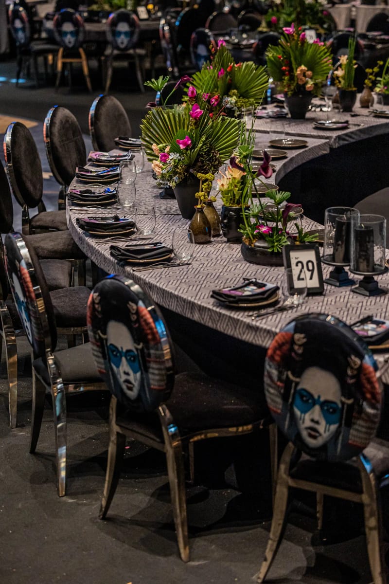 Curved table with black chairs whose backs carry illustrated portraits, tropical plants, pink flowers and a table number