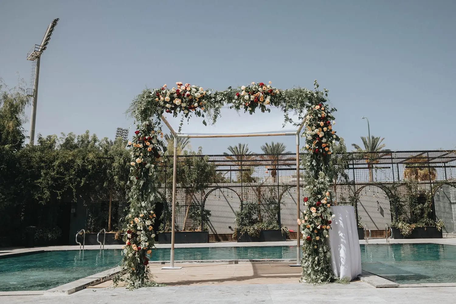 Arche florale rectangulaire de roses et de verdure au bord d'un bassin, avec palmiers et projecteur au fond