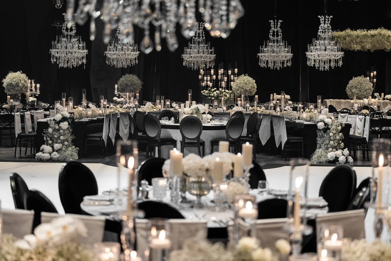 Lustres en cristal au-dessus de tables dressées avec bougies, fleurs blanches et chaises noires dans une salle sombre