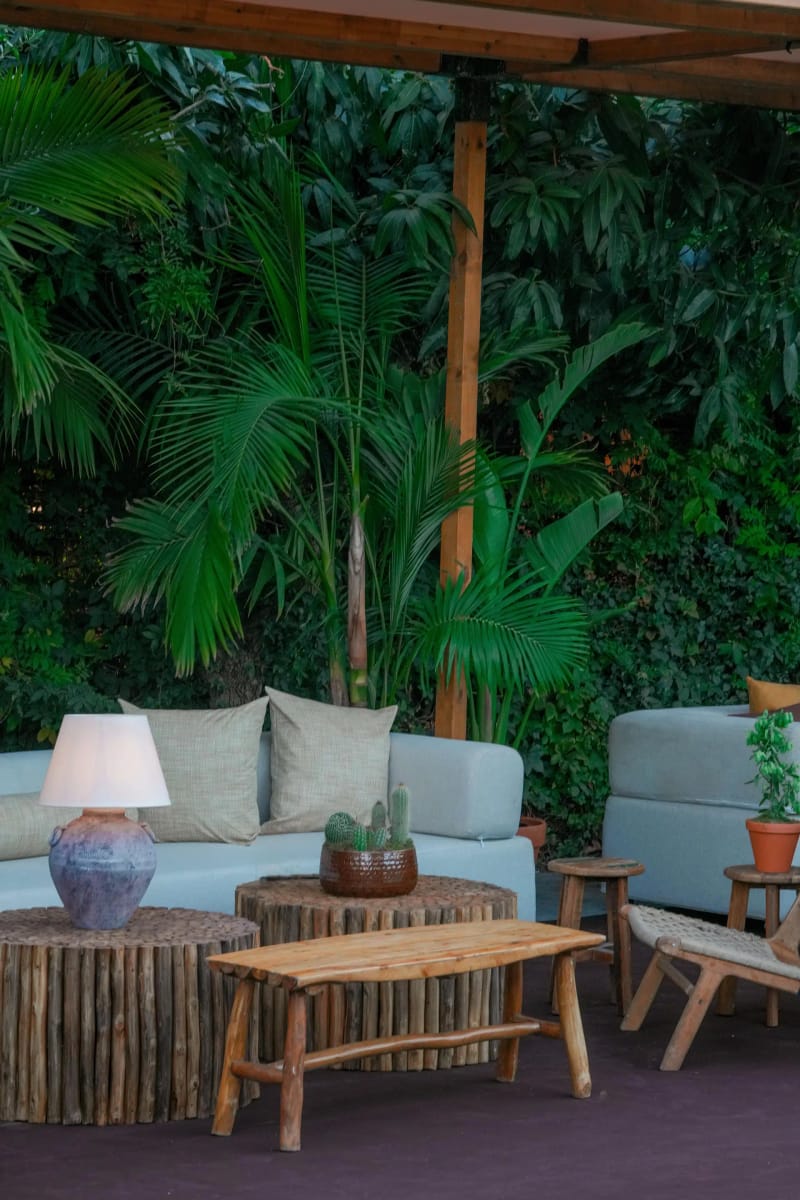 Lounge corner under a timber pergola with pale blue sofas, log-slice tables, a table lamp and palms against a green wall
