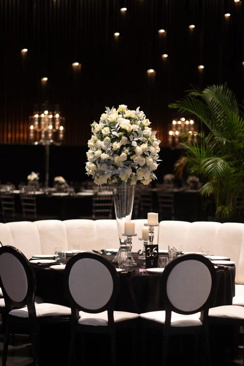 Tall white floral arrangement on a round table with black chairs, a curved cream banquette and a palm behind