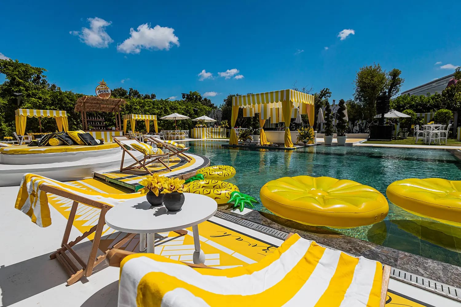 Round yellow floats in the pool, a deck chair with a striped towel and a small table, yellow cabanas in the distance