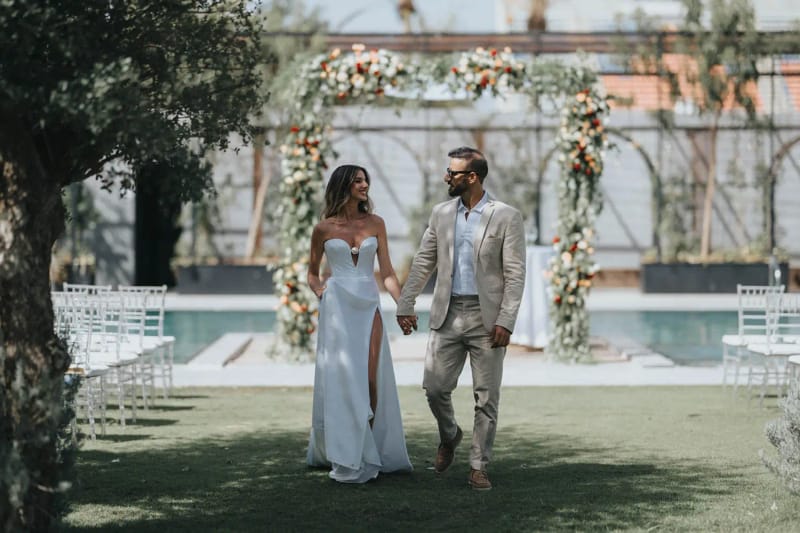 A couple walking hand in hand across the lawn toward a floral arch, with white chairs and a pool behind
