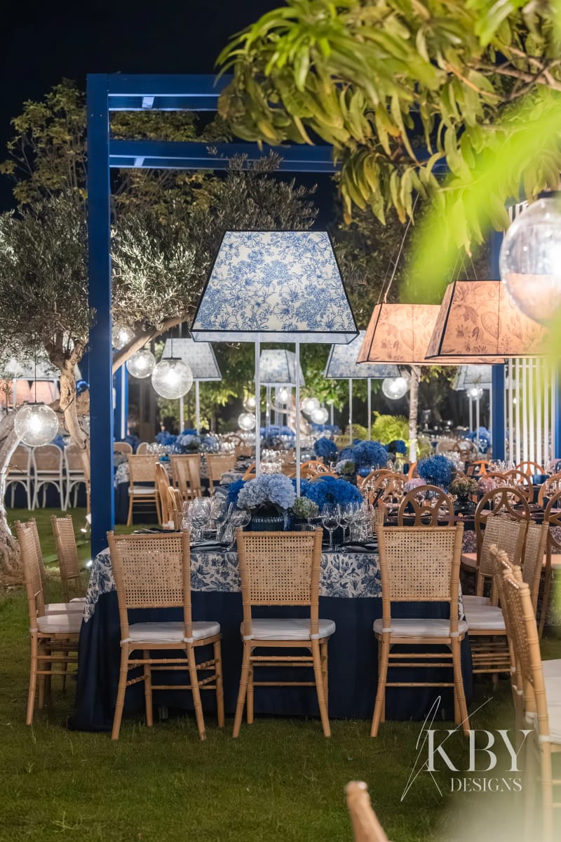 Cane-back chairs and a table with a blue and white patterned cloth on the lawn, with large lampshades hung from a blue frame