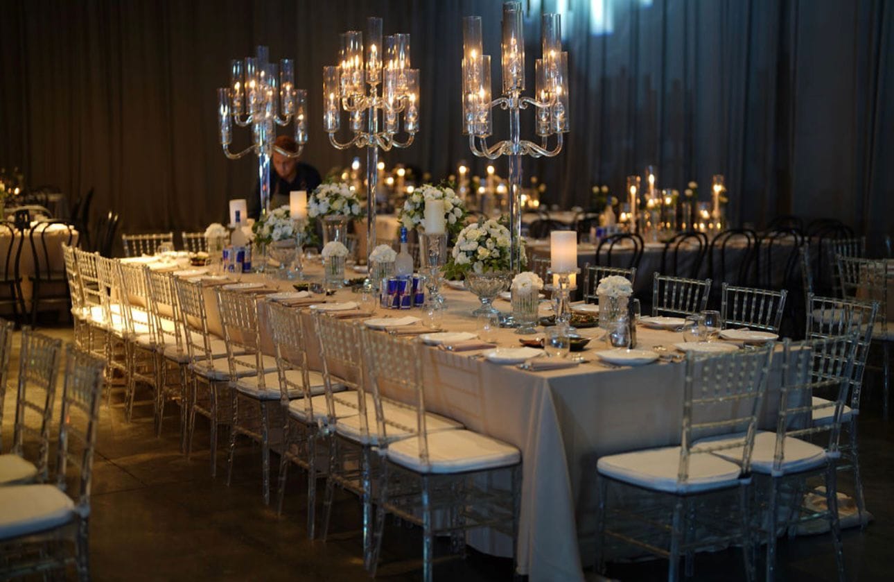 Longue table de banquet avec chaises transparentes, candélabres et fleurs blanches dans une salle aux rideaux sombres