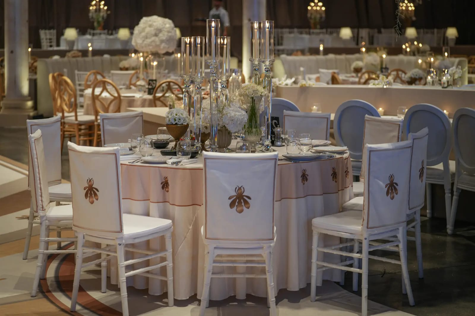 Table ronde à nappe crème et chaises blanches ornées d'un emblème doré brodé au dossier, avec des centres de table fleuris