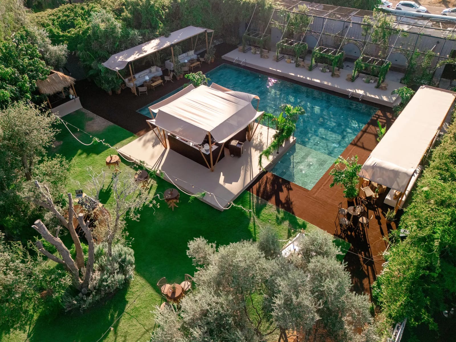 Aerial view of a pool with a fabric pavilion on a wooden deck and long canopies beside it, ringed by lawn and greenery