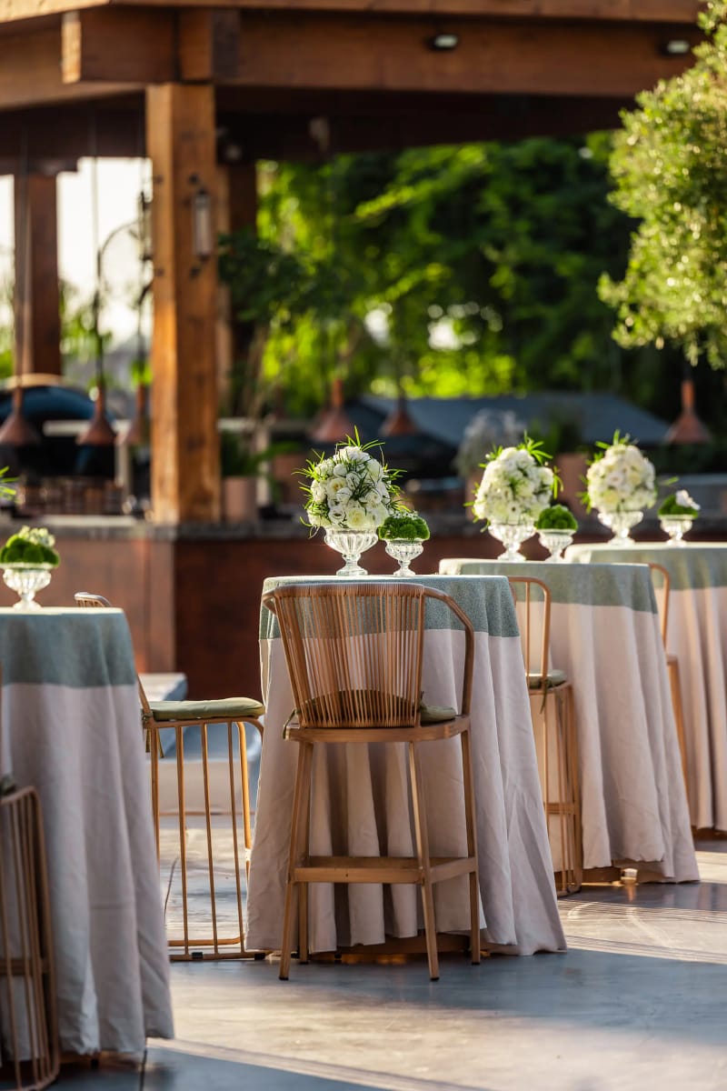 High cocktail tables with pale-green cloths and small floral arrangements, with bar stools under a wooden pergola