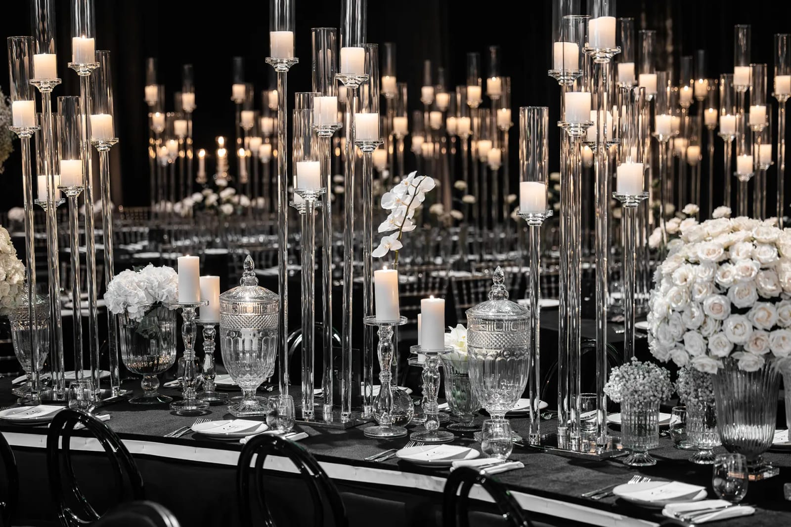 Ensemble de hauts cylindres en verre avec bougies, roses blanches et bonbonnière en cristal sur une table à nappe noire