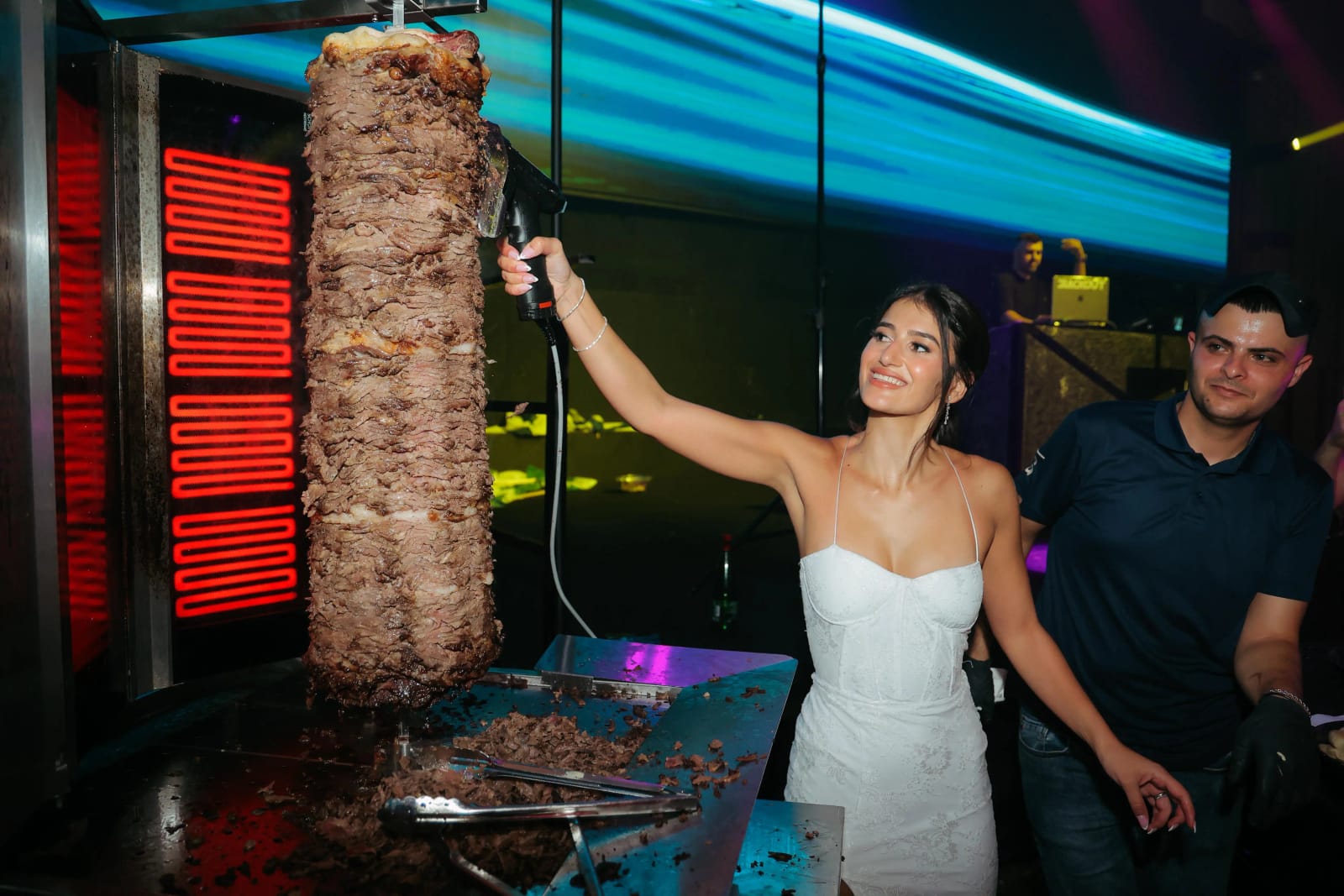 A bride in a white dress carving from a vertical meat spit at a colorfully lit food station, a man beside her