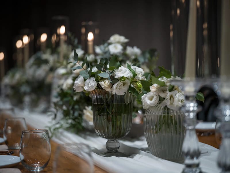 Close-up of a green glass vase with white flowers and foliage, lit candles blurred behind
