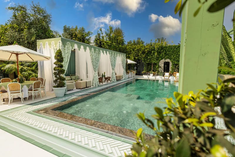 Vue en angle d'un coin de bassin avec parasol à franges, tables rondes, chaises tressées et topiaire devant un mur de lierre